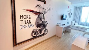 A modern hotel room with a large framed artwork of a bird on a motorcycle, a white sofa, a desk with a computer, and a window with curtains. - Beat Hotel Bangkok (Bangkok)