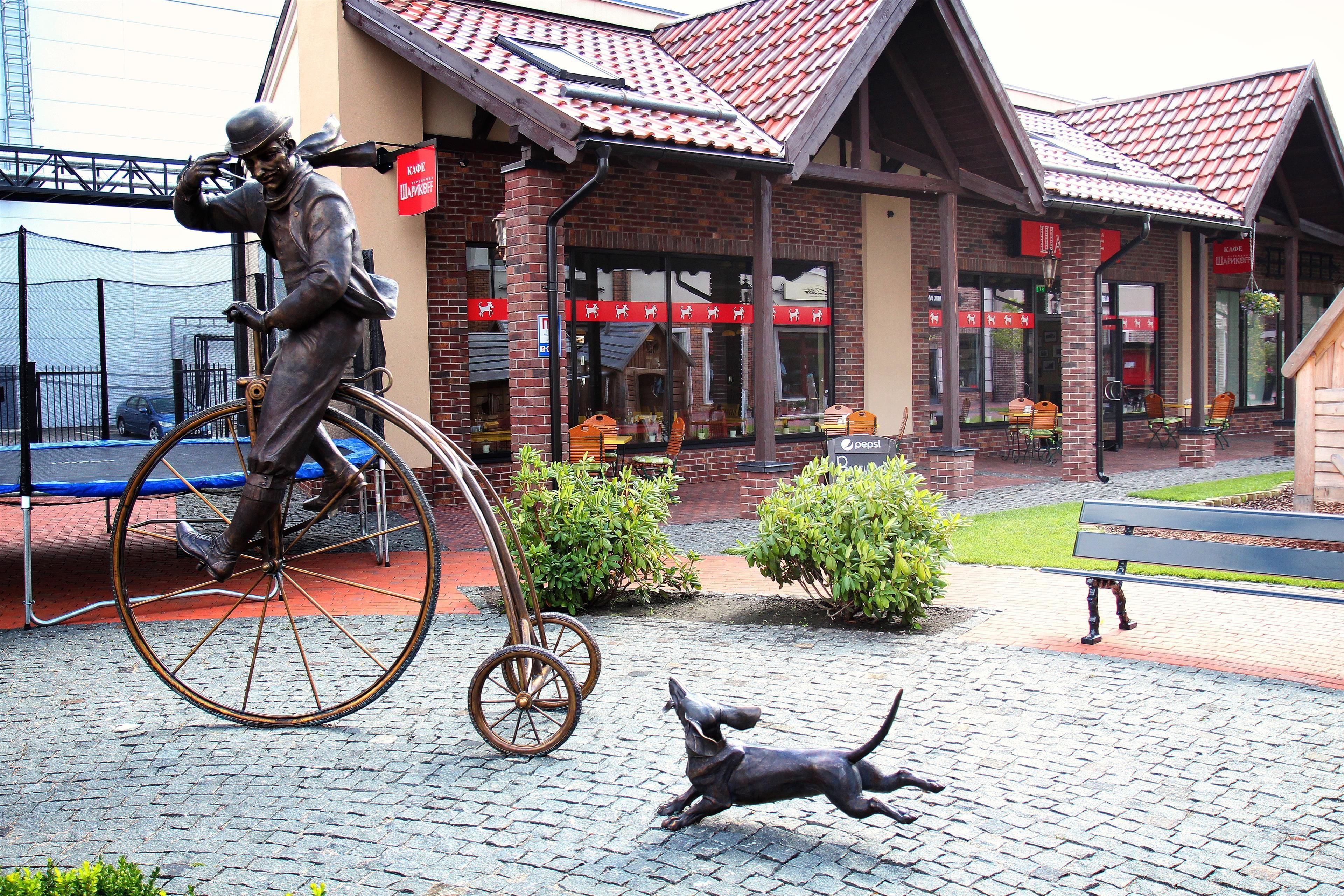 A statue of a man on a bicycle and a dog in front of a building with a red roof and brick walls.