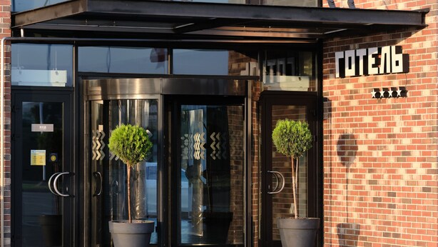 Entrada de hotel com porta de vidro, plantas em vasos e placa acima da porta.