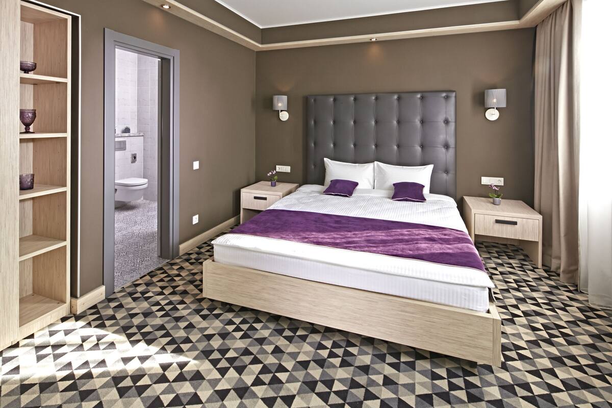 A modern bedroom with a large bed, two bedside tables, a built-in shelf unit, and a geometric patterned floor.