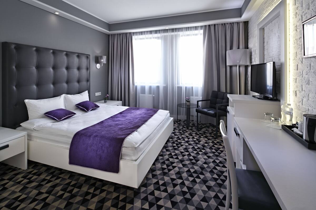 A modern hotel room with a large bed, a desk, a chair, a TV, and a window with curtains.