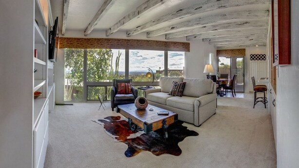 A spacious living room with a high ceiling, a sofa, a coffee table, and a large window offering a view of the outdoors. - Mission Hill Casitas (Tucson)