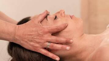 A person receiving a massage with hands on their head, in a room with a plain wall and a white towel.