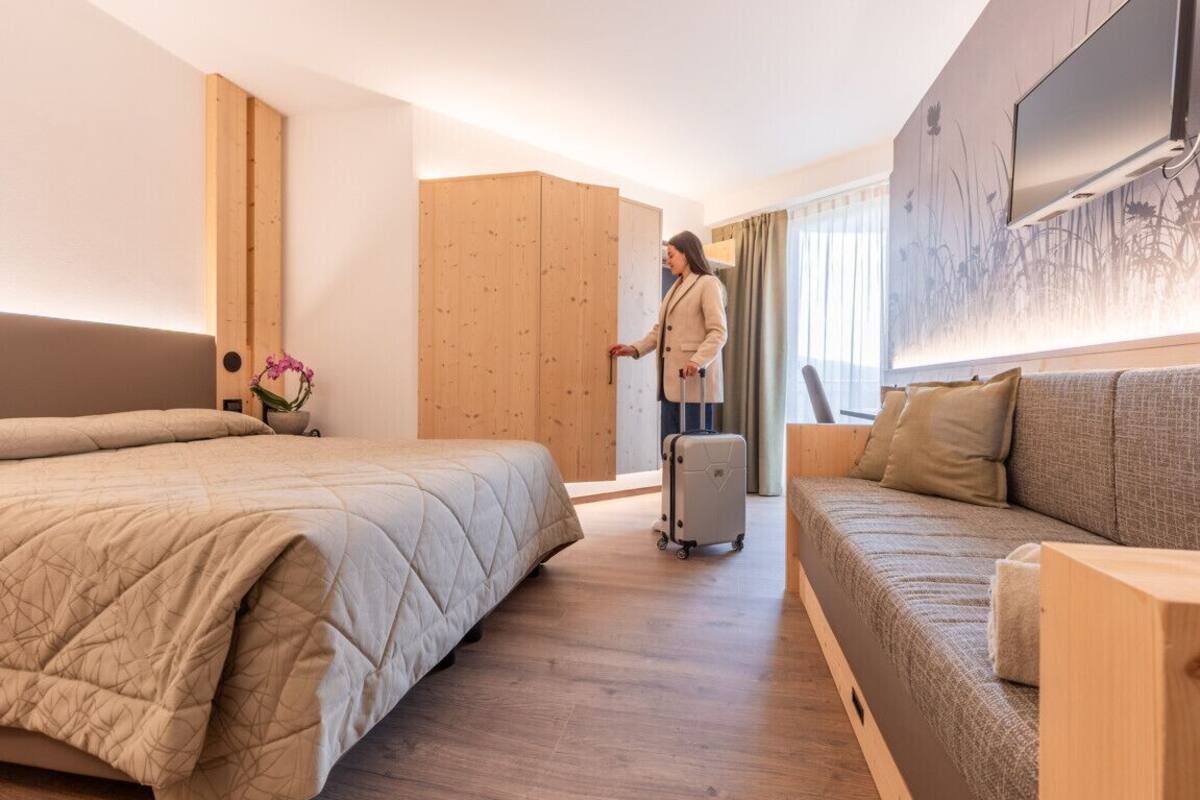 Une chambre d’hôtel avec un lit, un canapé, une armoire en bois, une valise et un téléviseur fixé au mur.