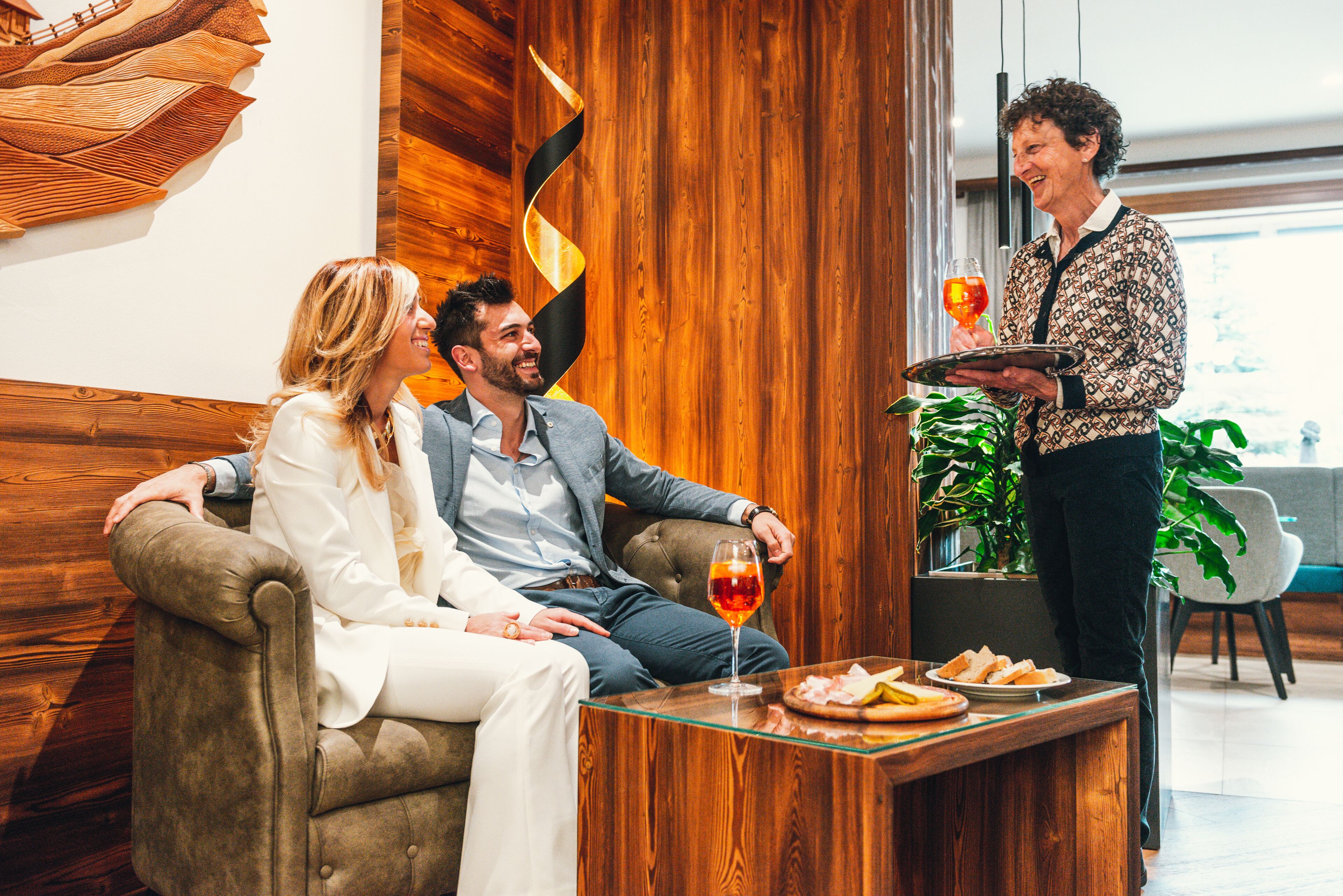 Ein Holzinterieur mit einer Couch, einem Tisch mit Speisen und einem Weinglas sowie einer Person, die bedient.