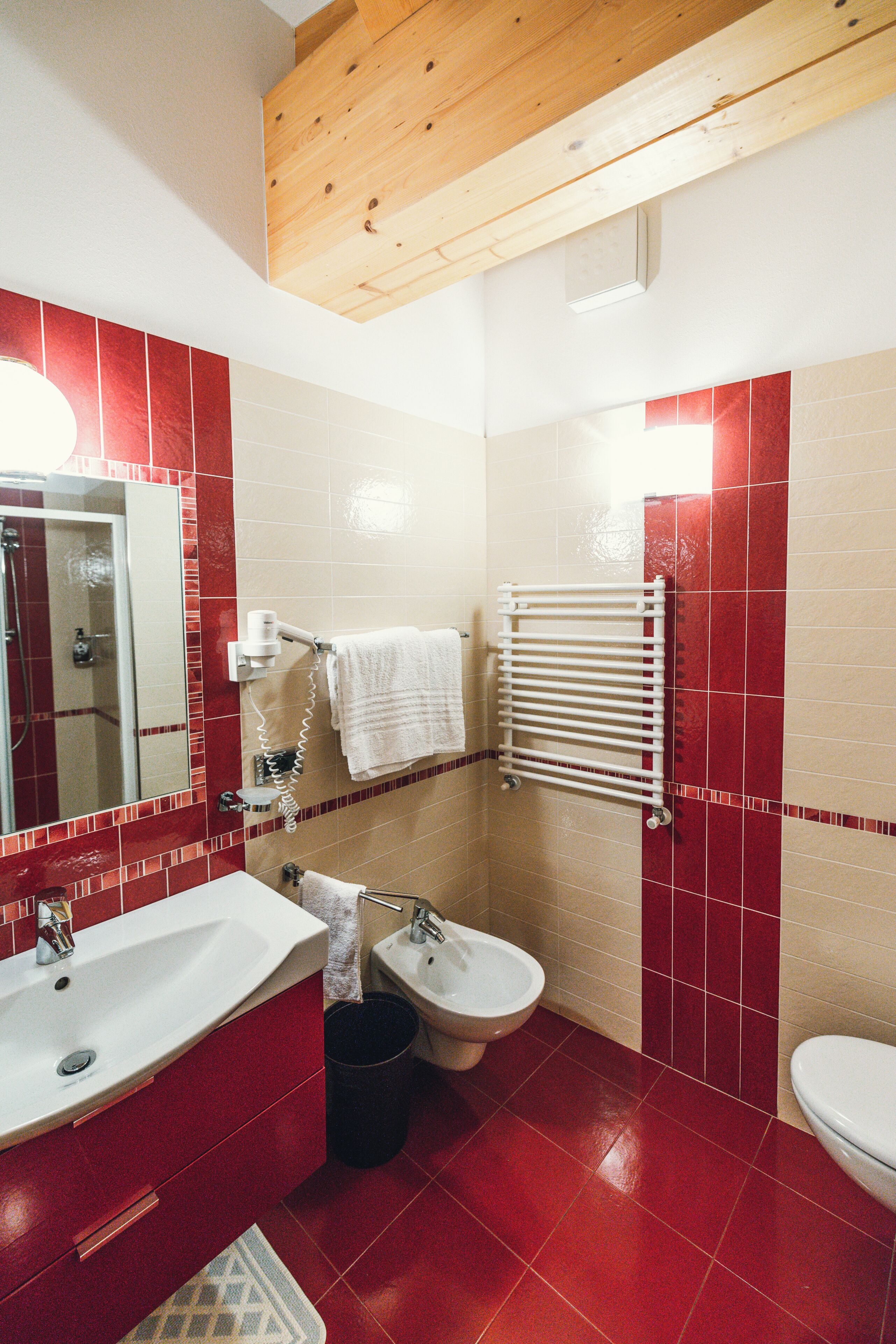 A bathroom with a sink, bidet, and toilet, red and beige tiles, a towel rack, and a wooden ceiling beam.