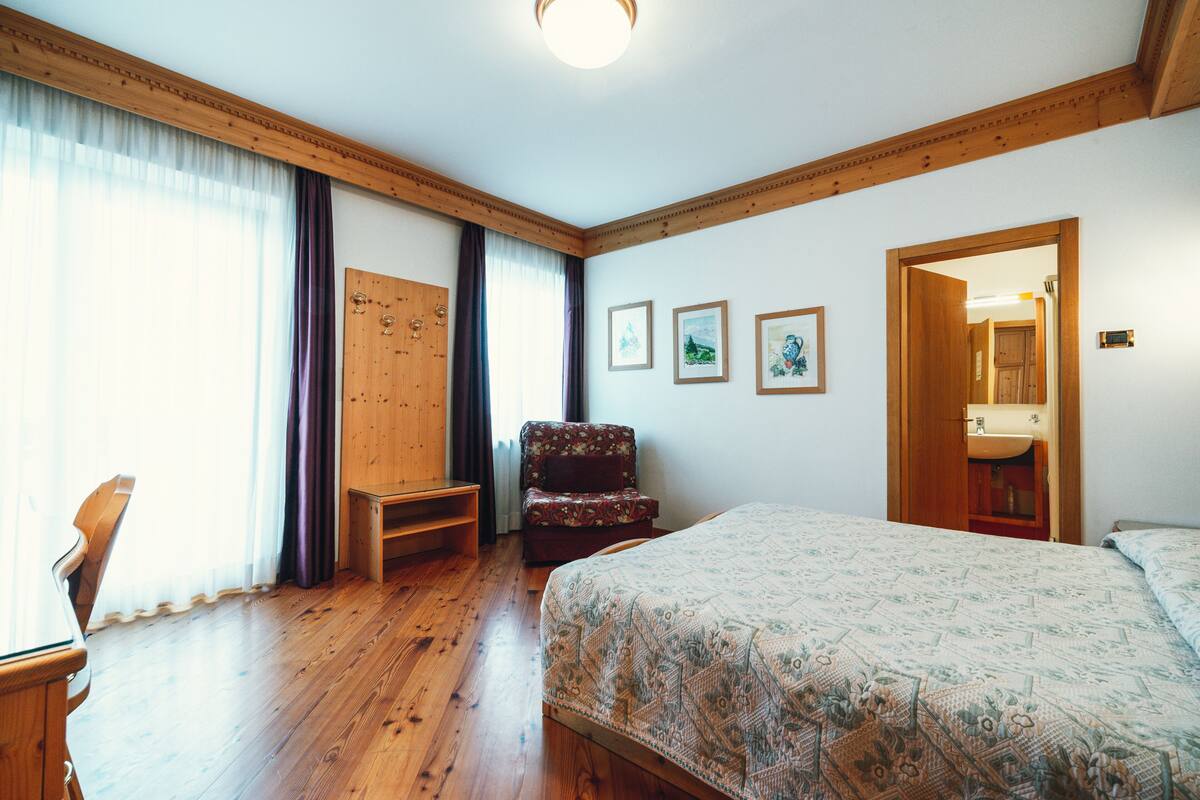 Une chambre avec un plancher en bois, un lit, une chaise, un bureau et une porte donnant sur une salle de bain.