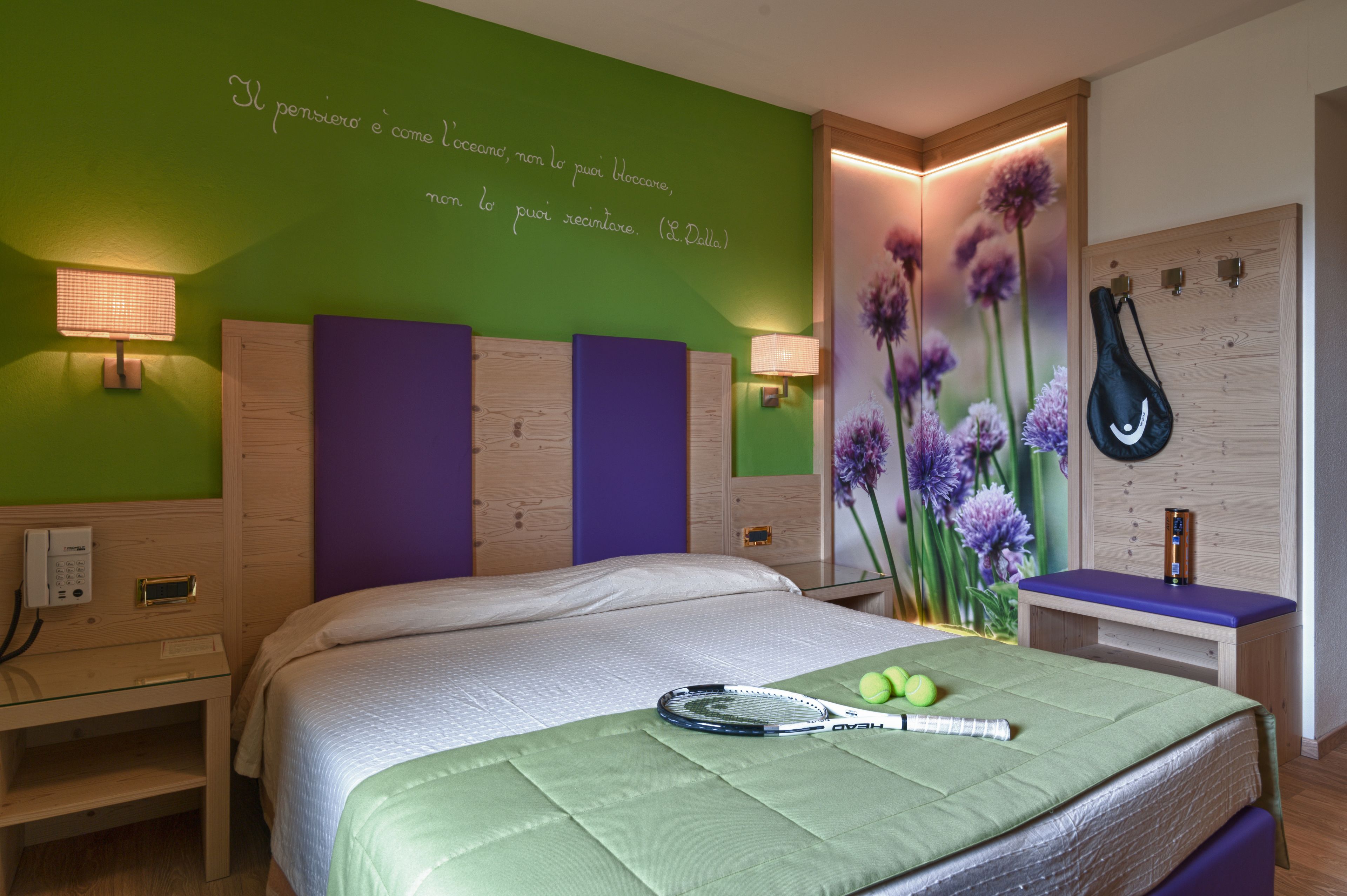 Habitación de hotel con pared verde, una cama con cubrecama verde, una raqueta de tenis y pelotas de tenis sobre la cama.