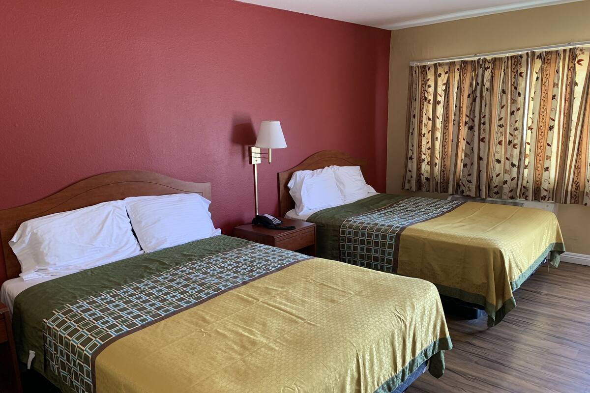 A hotel room with two beds, a nightstand with a lamp, and a window with floral curtains.