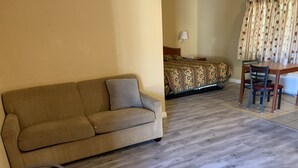 A hotel room with a sofa, a bed with a floral bedspread, a table with chairs, and a lamp. - Desert Inn Motel (Corona)