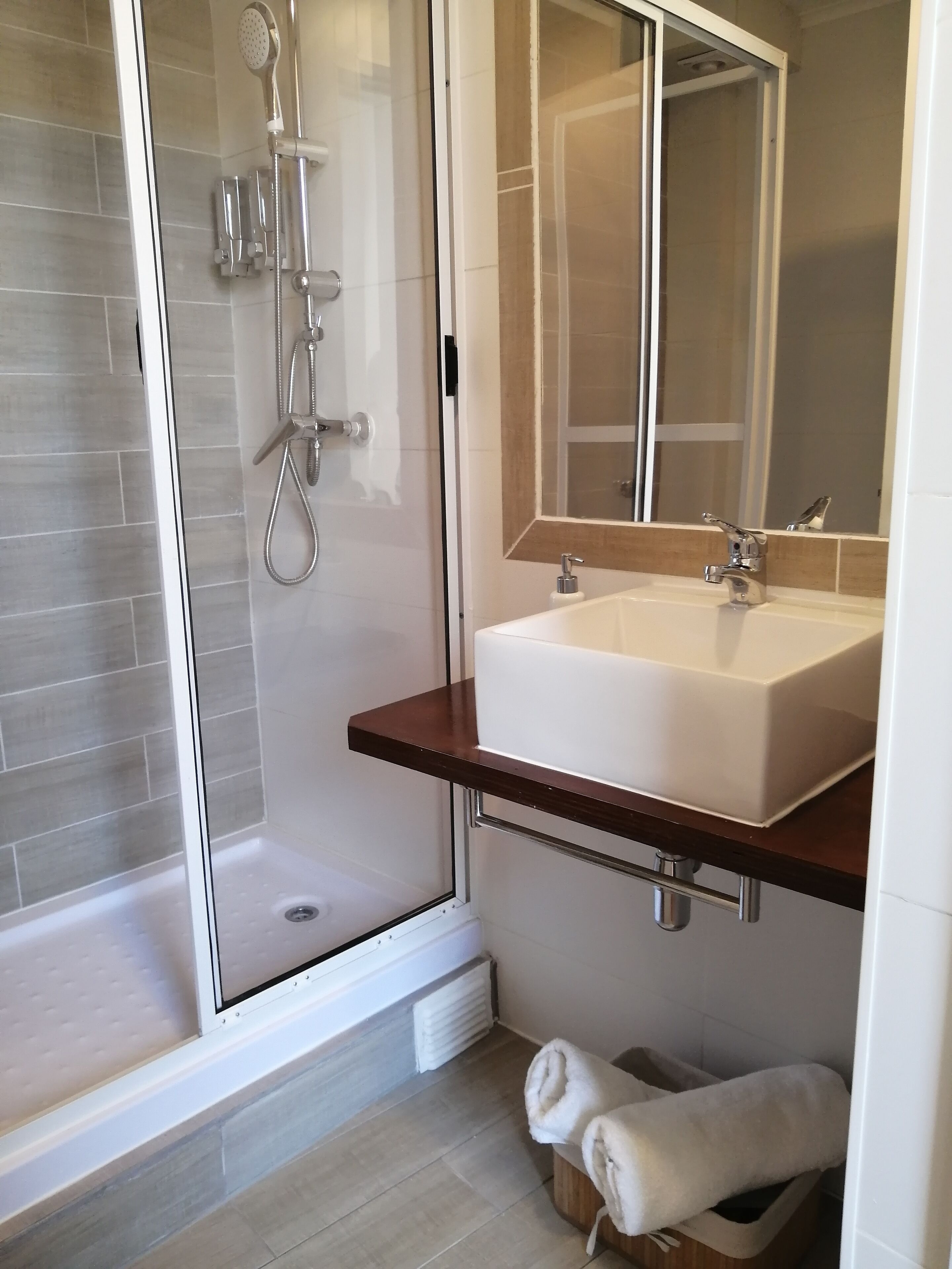 Ein modernes Badezimmer mit einer ein Glaswand trennenden Dusche, einem weißen Waschbecken mit Holzablage und einem Spiegel darüber.