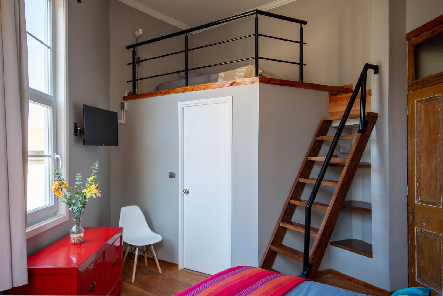 Un dormitorio con una cama individual sobre otra, una escalera de madera, un escritorio con silla, un aparador rojo y una ventana con flores.