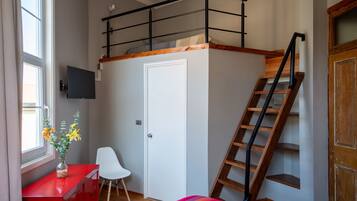 Un dormitorio con una cama individual sobre otra, una escalera de madera, un escritorio con silla, un aparador rojo y una ventana con flores.