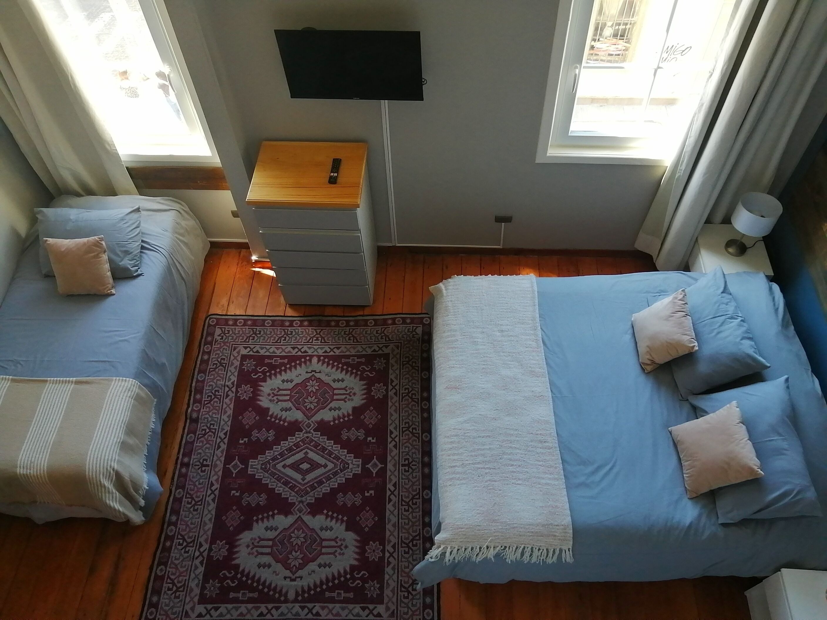 Un dormitorio con una cama, una mesita de noche de madera, un televisor montado en la pared y una ventana con cortinas.
