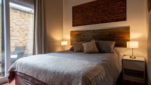 Quarto com cama grande, duas luminárias de cabeceira, painel de madeira e vista para uma parede de tijolos pela janela.