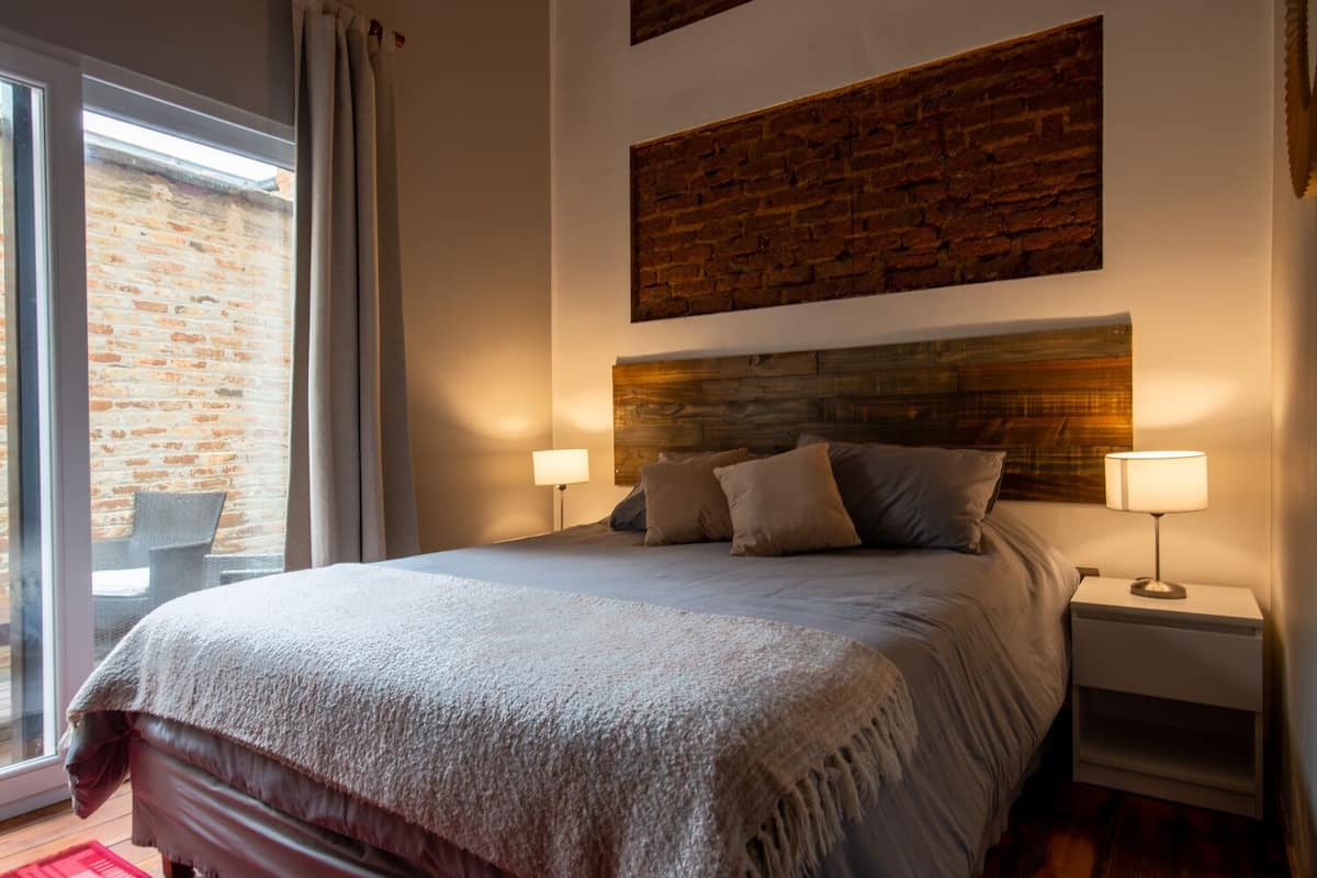 A bedroom with a large bed, two bedside lamps, a wooden headboard, and a view of a brick wall through a window.