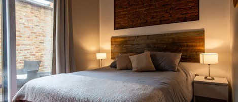 A bedroom with a large bed, two bedside lamps, a wooden headboard, and a view of a brick wall through a window.