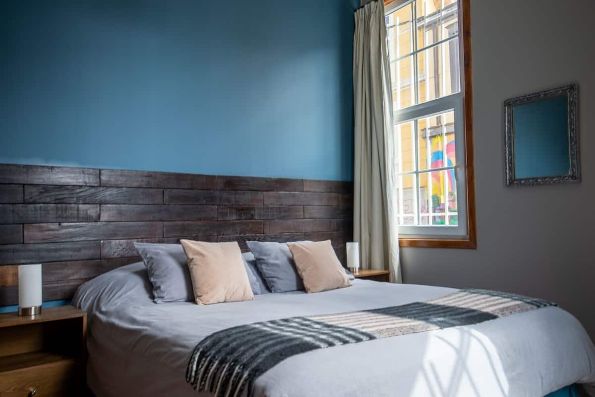 A bedroom with a wooden headboard, a large bed with multiple pillows, a bedside table with a lamp, and a window with curtains.