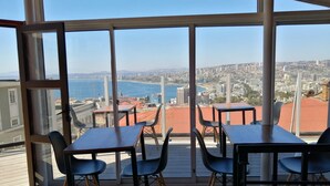 Uma sala de jantar com janelas amplas, mesas e cadeiras, com vista para uma paisagem urbana e um corpo de água.