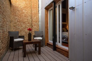 Un pequeño patio con sillas de mimbre, una mesa, un muro de ladrillo y un ventanal corredero que da a un dormitorio.