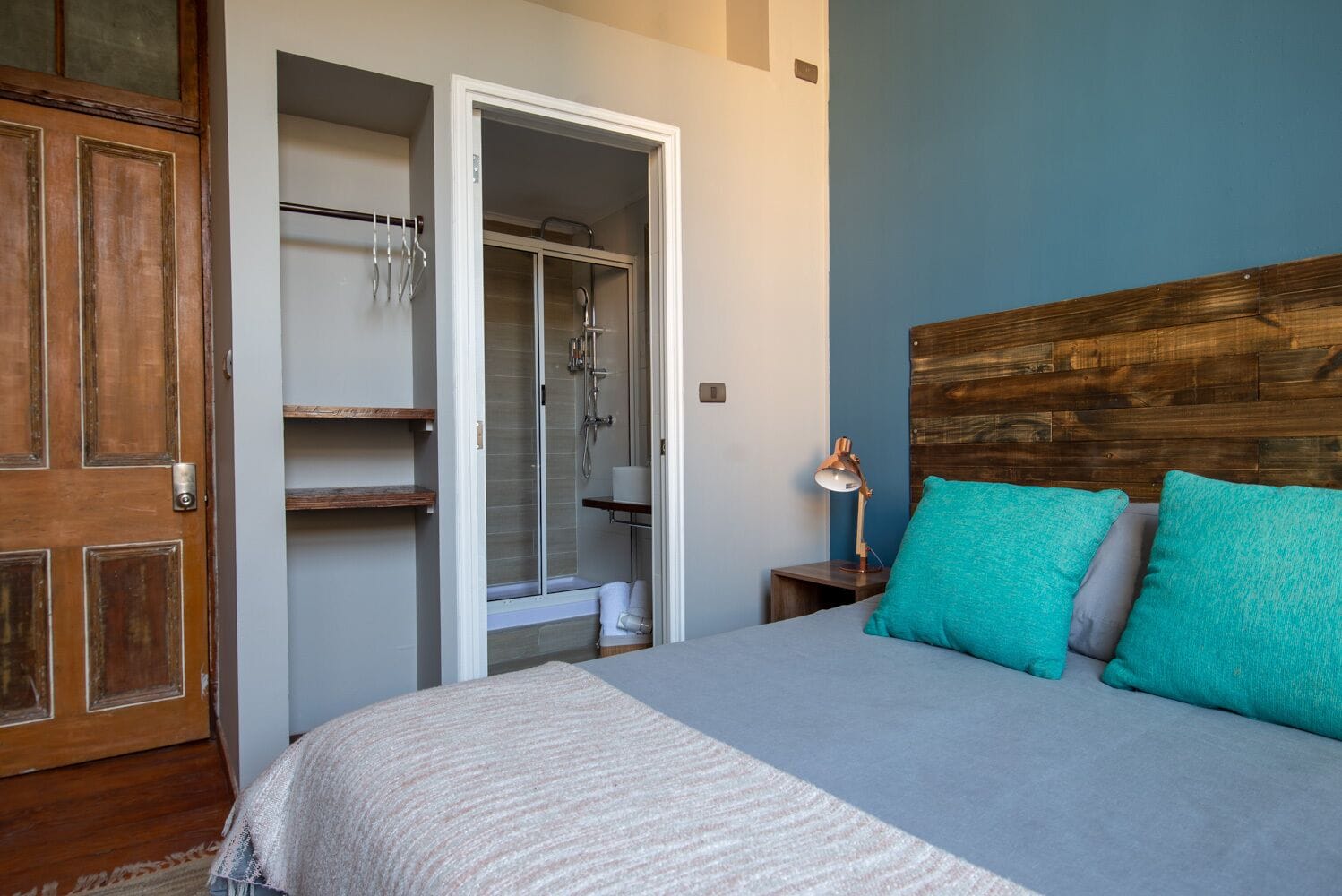 A bedroom with a bed, a wooden headboard, a nightstand with a lamp, a closet with a shower, and a door.