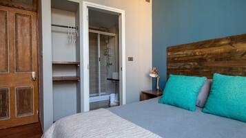 Un dormitorio con cama, cabecera de madera, mesita de noche con lámpara, armario con ducha y puerta.