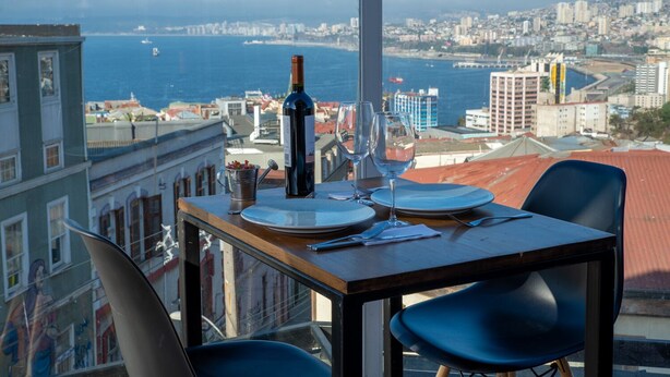 Mesa de jantar posta com pratos, copos de vinho e uma garrafa, com vista para um horizonte urbano com edifícios e um corpo de água.