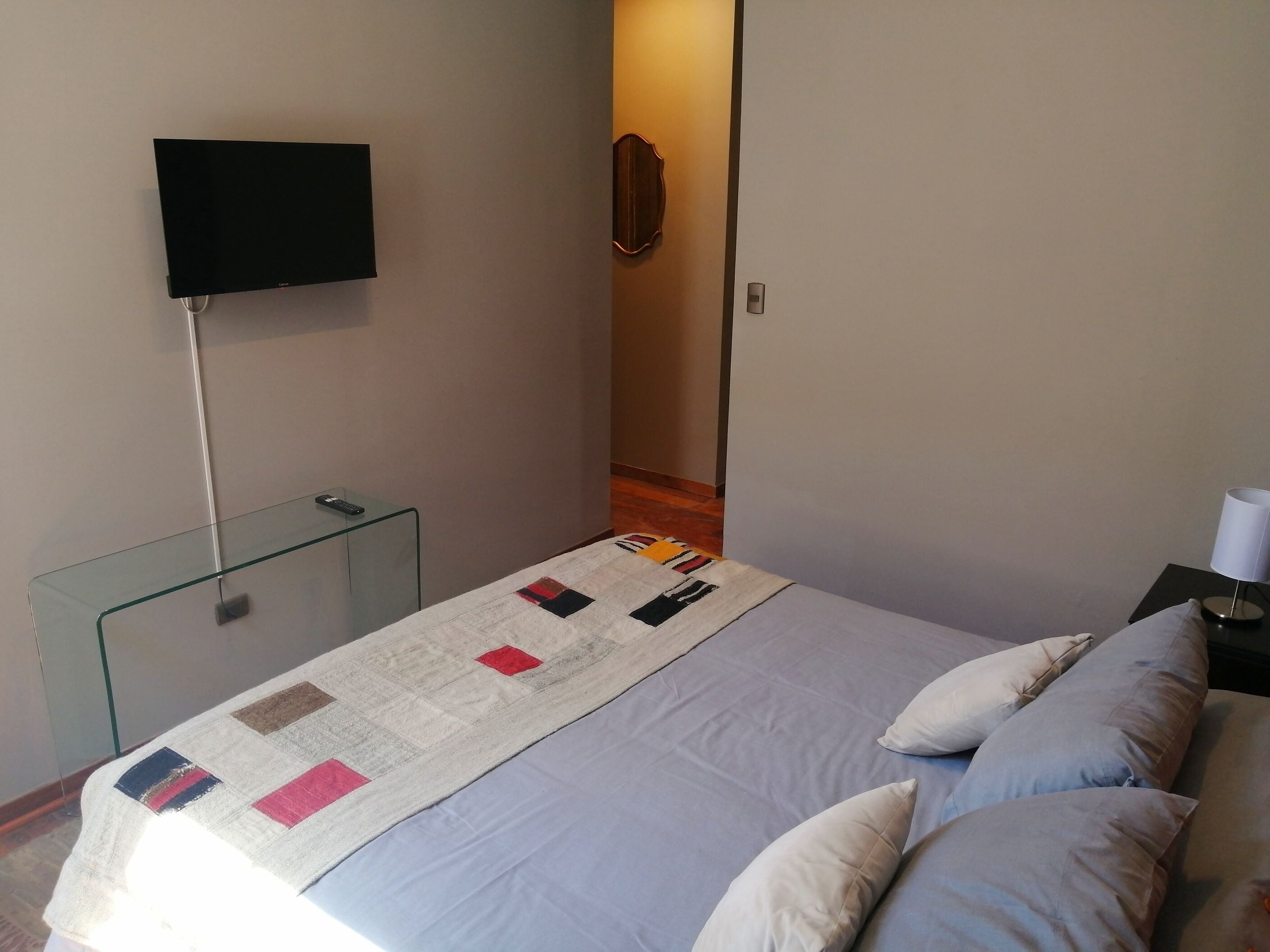 A bedroom with a bed, a TV mounted on the wall, a bedside table, and a lamp.
