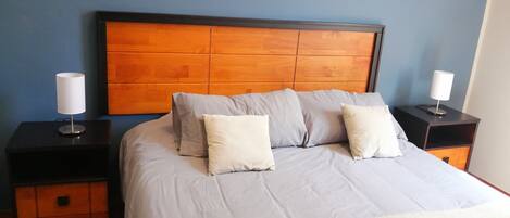 Un dormitorio con cabecera de madera, colcha gris y dos mesitas de noche con lámparas.