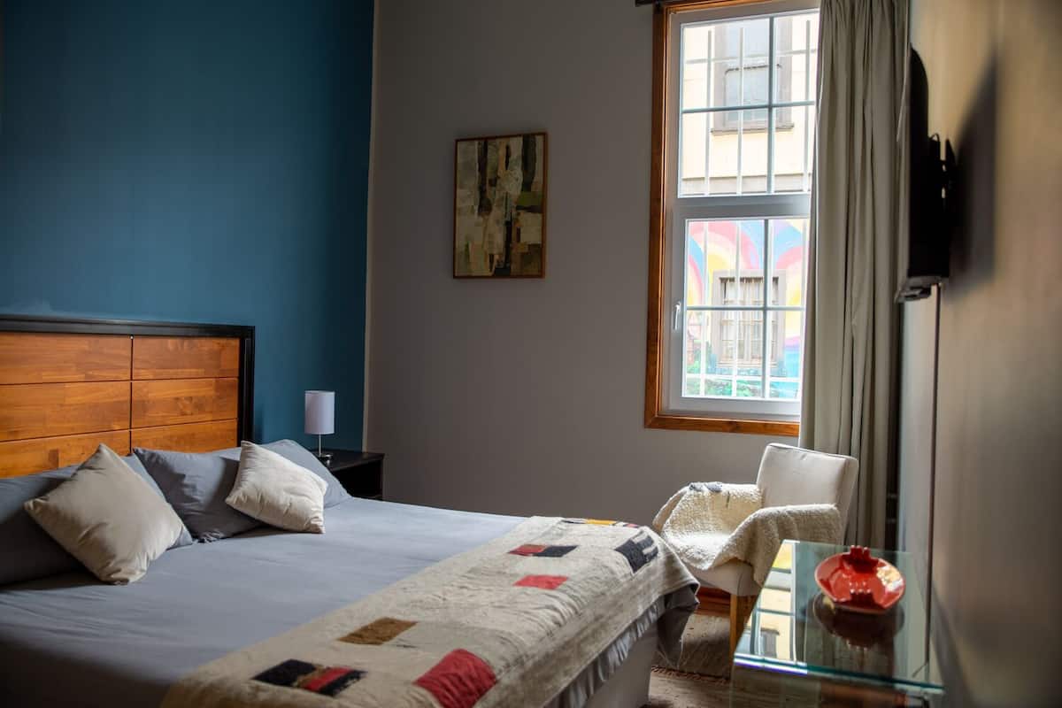 A bedroom with a bed, a chair, a nightstand, a window, and a painting on the wall.