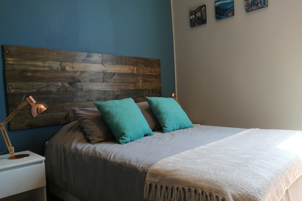 A bed with a wooden headboard, two teal pillows, a white throw blanket, and a bedside lamp.