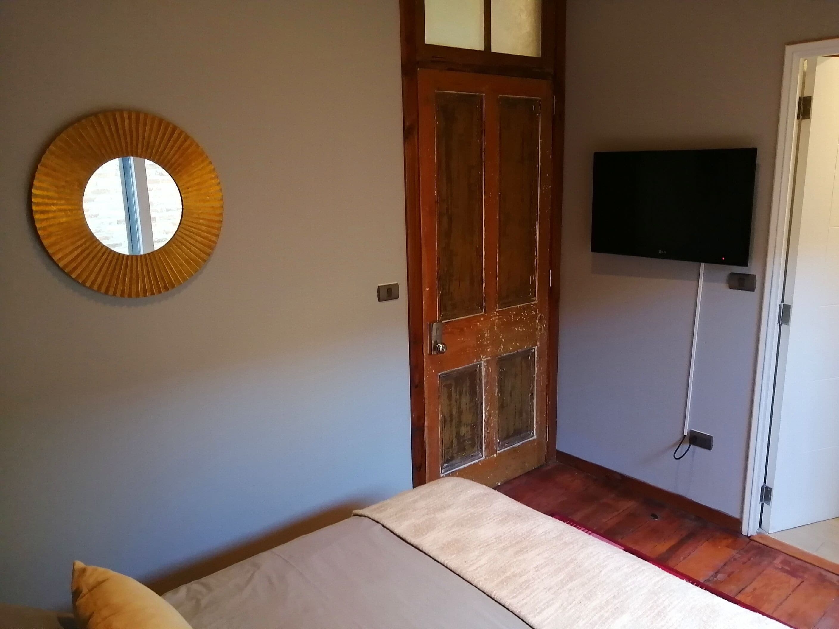 A bedroom with a bed, a wooden door, a television mounted on the wall, and a round mirror.