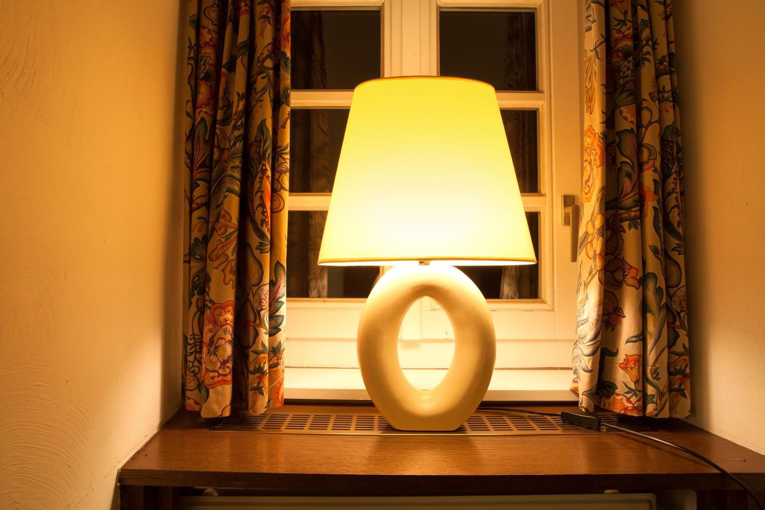 A table lamp with a unique circular base and a yellow lampshade, positioned on a wooden table by a window with floral curtains.