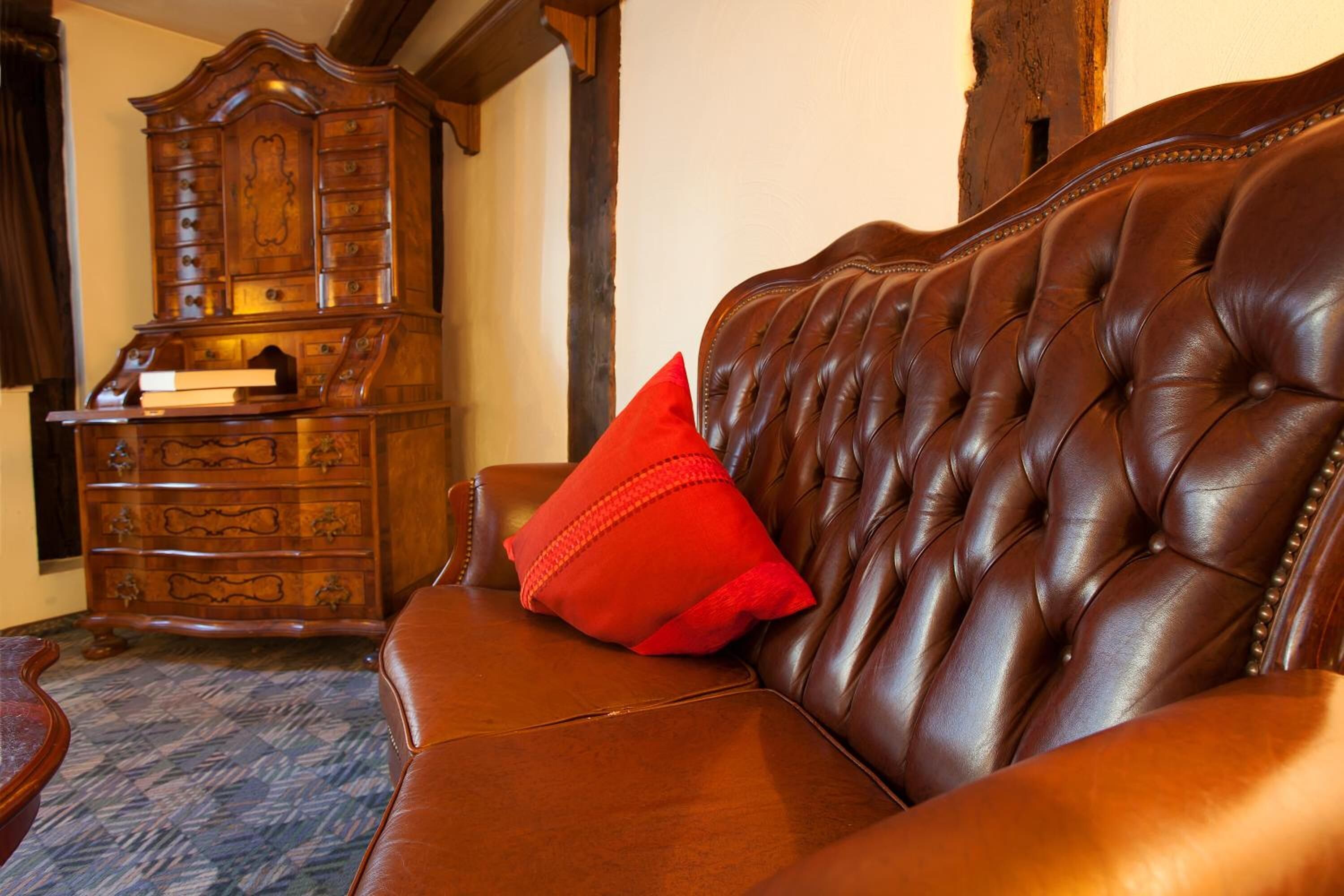 A brown leather sofa with a red pillow, a wooden cabinet with drawers, and a patterned carpet.
