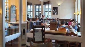 Vista interna de um restaurante com vigas de madeira no teto, janelas amplas e clientes jantando.