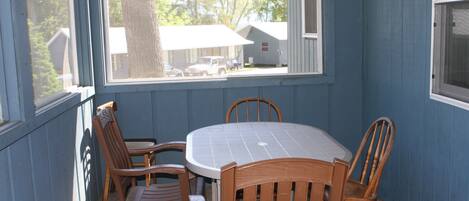 Une terrasse couverte avec une table ronde et quatre chaises en bois, donnant sur un quartier résidentiel avec des maisons et un véhicule garé.