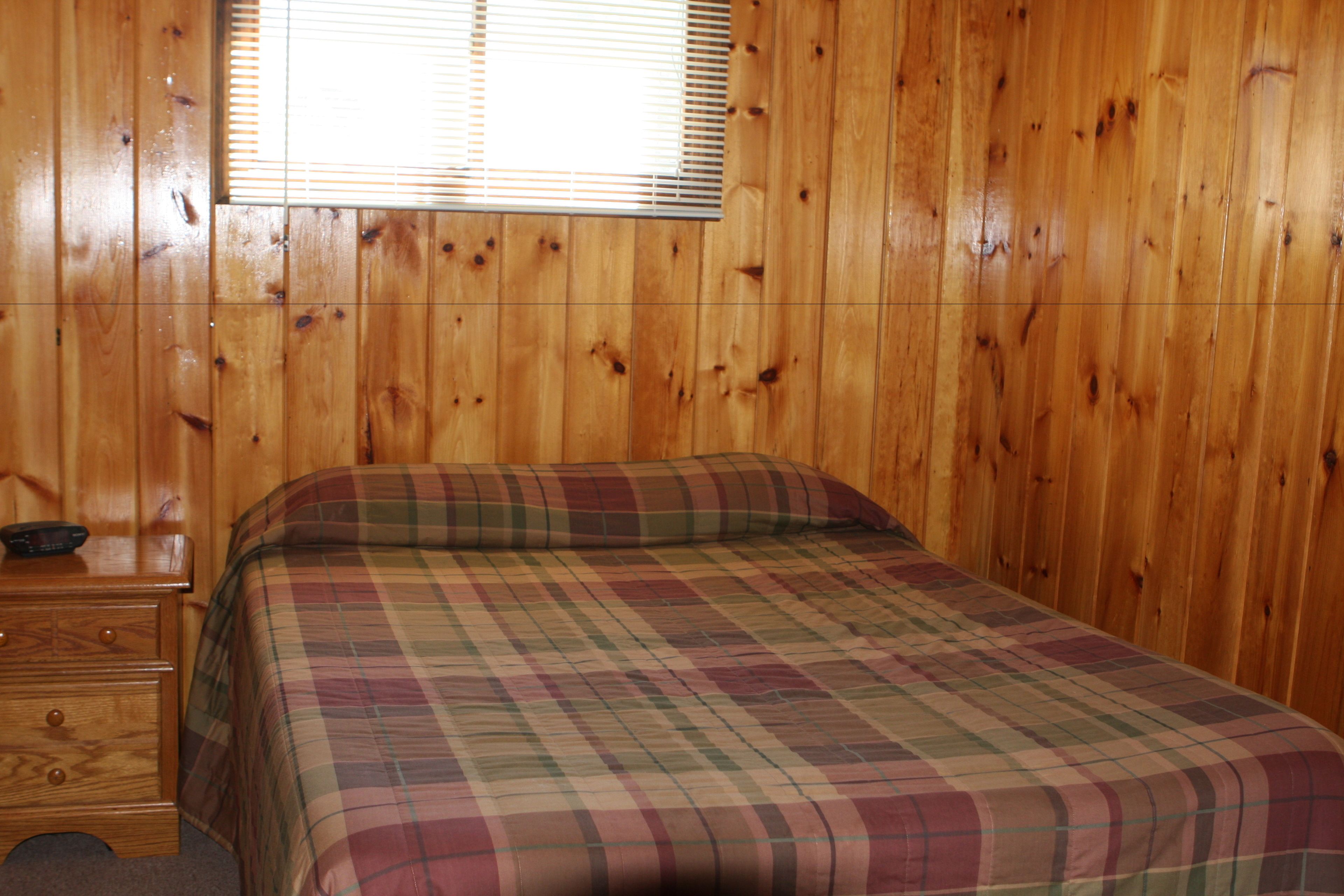 Een kamer met houten wanden, een bed met een geruit sprei, een nachtkastje van hout met lades en een raam met jaloezieën.