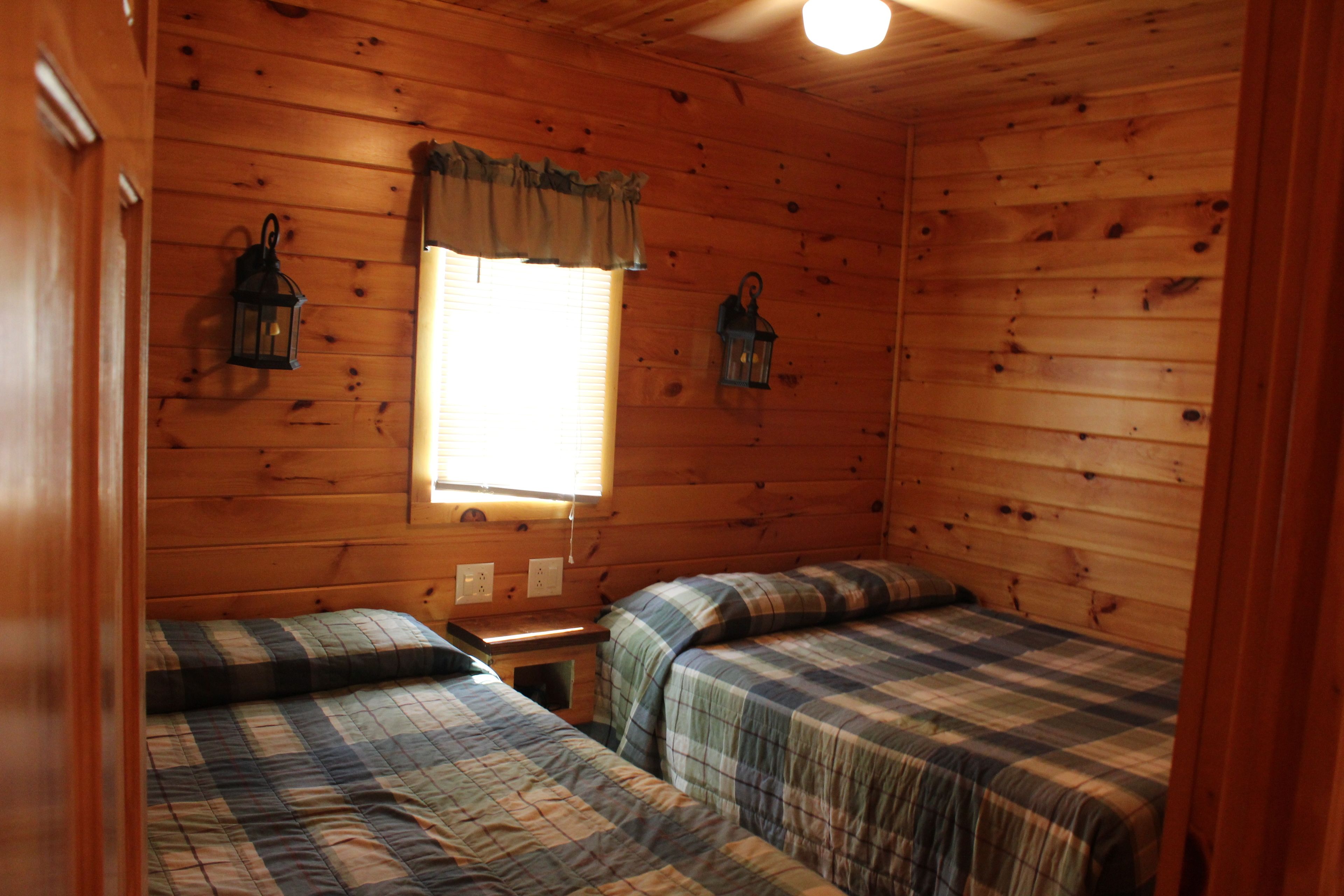 A cabin-style room with two beds, wooden walls, a window with curtains, and two wall-mounted lanterns.