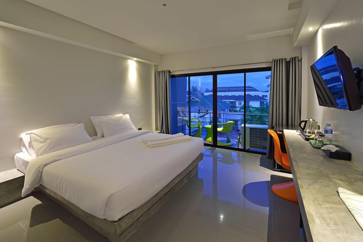 A modern hotel room with a large bed, a desk with a flat-screen TV, and a balcony with a city view.