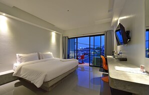 A modern hotel room with a large bed, a desk, a TV, and a view of the city at dusk. - Riverside Hotel (Krabi)