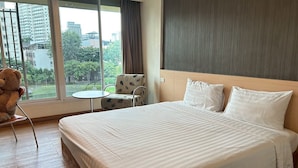A hotel room with a large bed, a small round table, a chair, and a view of the city through large windows. - S3 Residence Park (Bangkok)