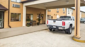Una camioneta pickup blanca estacionada bajo una estructura cubierta, con un banco y plantas en macetas cerca.