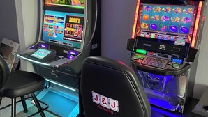 Dos máquinas tragamonedas con el logotipo de J&J Gaming, un televisor sobre la máquina izquierda y una silla al frente.
