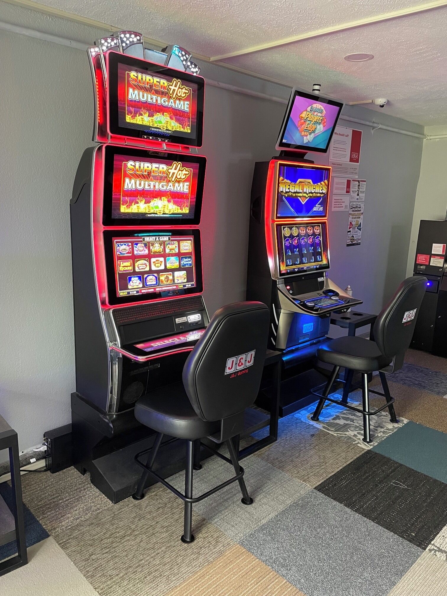 Two gaming machines with illuminated screens, a selection of chairs, and a wall-mounted sign.