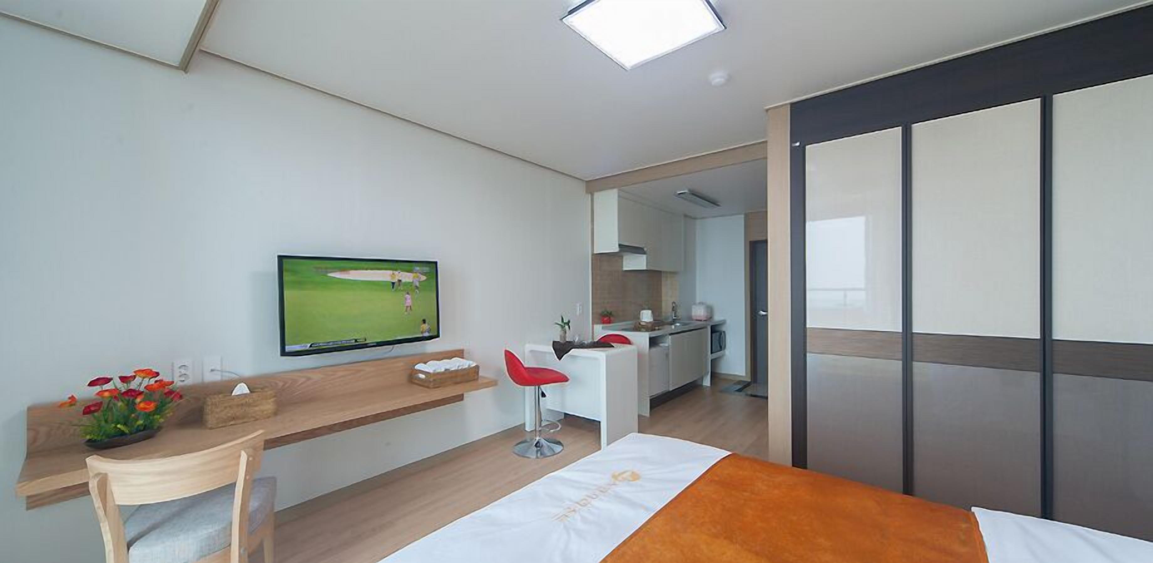A modern hotel room with a bed, a desk with a chair, a TV, and a kitchenette.