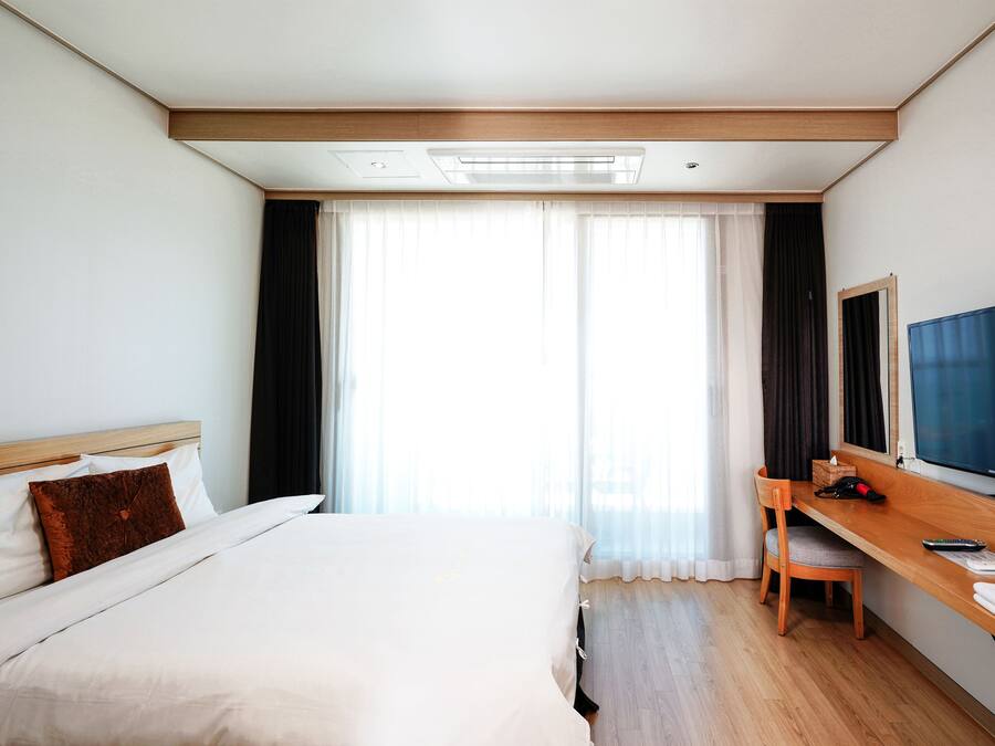 A hotel room with a bed, a desk with a chair, a television, and a large window with curtains.