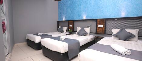 Habitación de hotel con tres camas, cada una con cabecero gris, ropa de cama blanca y una toalla enrollada sobre las camas.