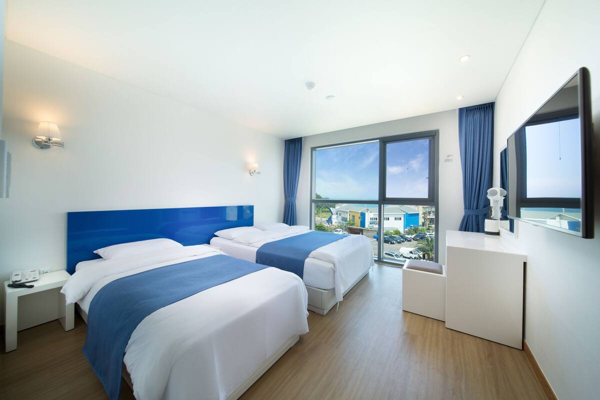 Hotellihuone, jossa on kaksi sänkyä, työpöytä, lipasto ja suuret ikkunat, joista avautuu näkymä kaupunkiin.