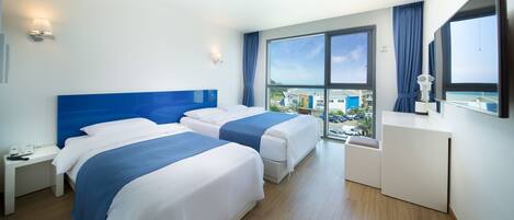 A hotel room with two beds, a desk, a dresser, and large windows offering a view of the city.