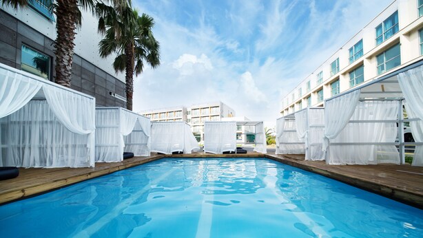 Piscina con toldos blancos y terraza de madera, rodeada de edificios modernos.
